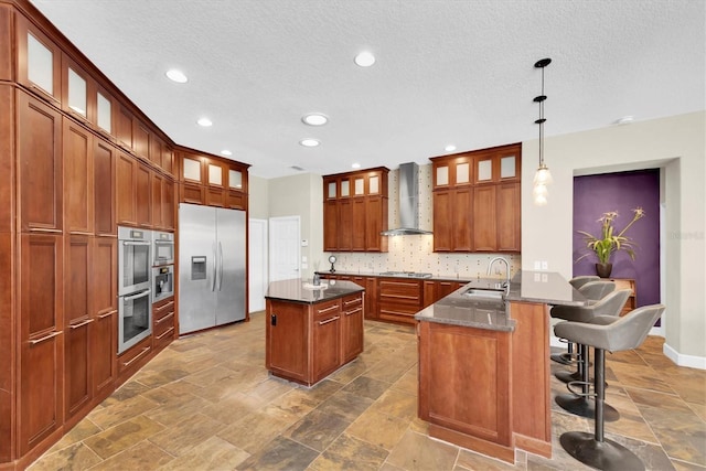 kitchen featuring a kitchen island with sink, a sink, stainless steel appliances, decorative backsplash, and wall chimney exhaust hood