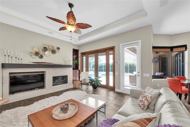 living room featuring stone tile flooring, a fireplace, ceiling fan, a raised ceiling, and crown molding