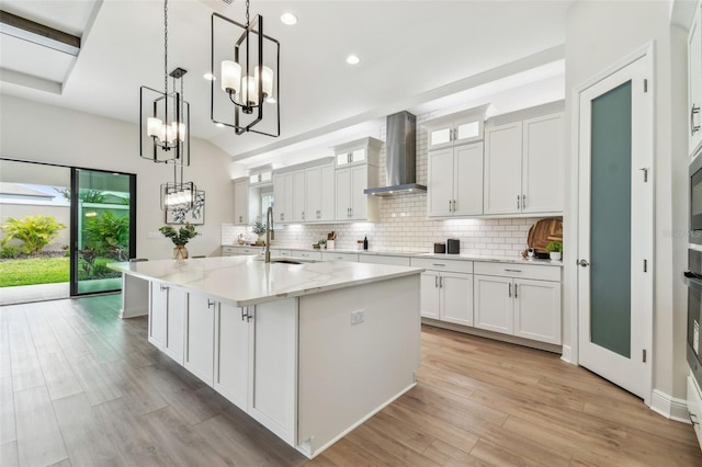 kitchen featuring an inviting chandelier, wall chimney exhaust hood, white cabinetry, a sink, and backsplash