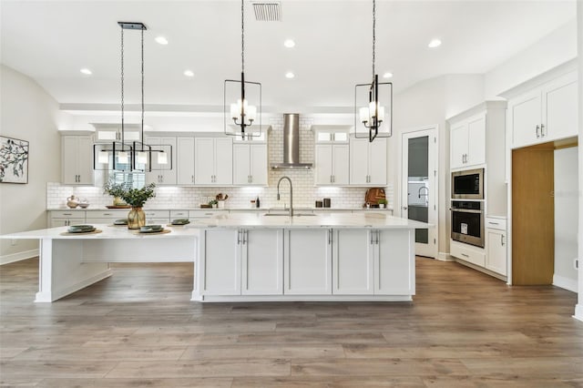 kitchen featuring an inviting chandelier, wall chimney exhaust hood, stainless steel oven, a large island with sink, and a sink