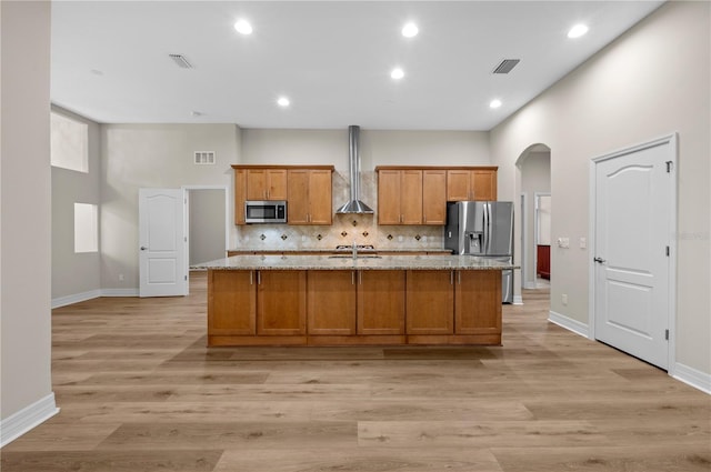 kitchen with arched walkways, an island with sink, stainless steel appliances, wall chimney range hood, and brown cabinets
