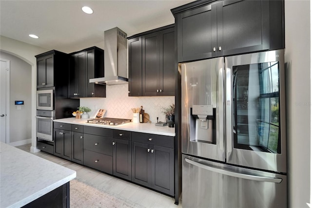 kitchen featuring tasteful backsplash, wall chimney range hood, light countertops, appliances with stainless steel finishes, and arched walkways
