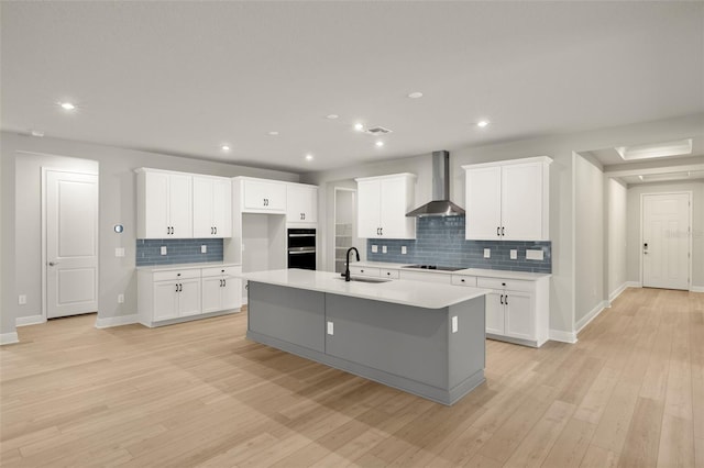 kitchen with wall chimney range hood, a sink, a kitchen island with sink, black electric cooktop, and tasteful backsplash