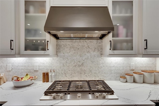 kitchen with glass insert cabinets, decorative backsplash, light stone countertops, stainless steel gas stovetop, and wall chimney exhaust hood