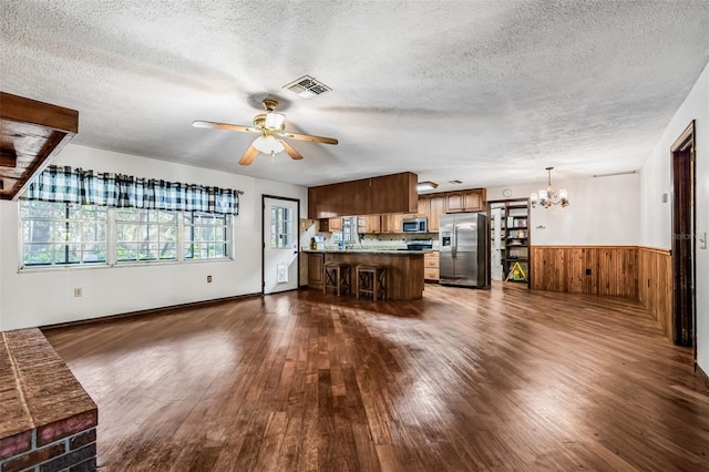 unfurnished living room with dark wood-style floors, a textured ceiling, a wainscoted wall, visible vents, and ceiling fan with notable chandelier