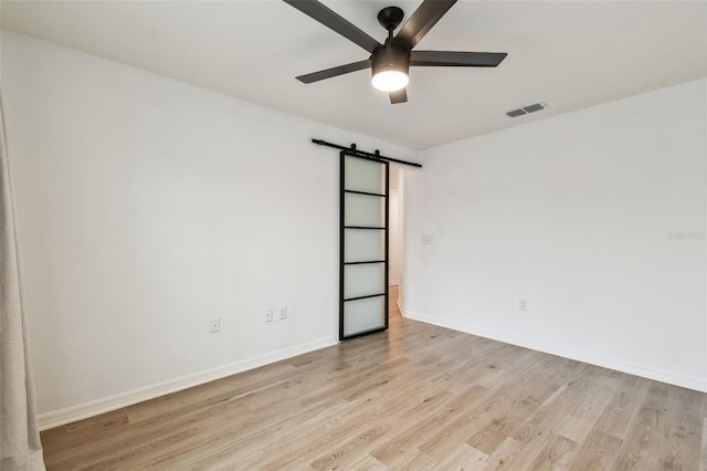 empty room with a barn door, light wood-type flooring, visible vents, ceiling fan, and baseboards