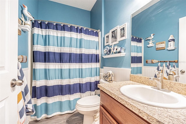 bathroom with toilet, wood finished floors, a shower with curtain, a wainscoted wall, and vanity
