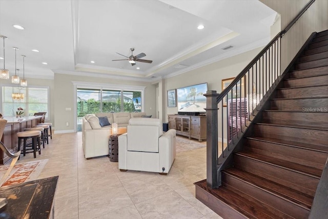 living room with stairway, crown molding, visible vents, a raised ceiling, and ceiling fan with notable chandelier