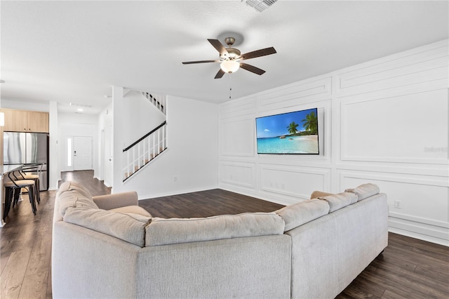 living room with a decorative wall, ceiling fan, stairway, and dark wood-style floors