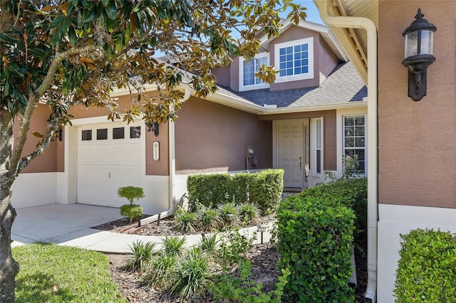 entrance to property with a shingled roof, an attached garage, driveway, and stucco siding