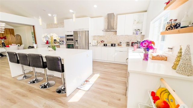 kitchen featuring open shelves, light wood-type flooring, stainless steel appliances, a kitchen bar, and wall chimney exhaust hood