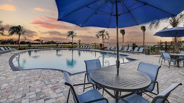 pool at dusk with a patio area, outdoor dining space, fence, and a community pool
