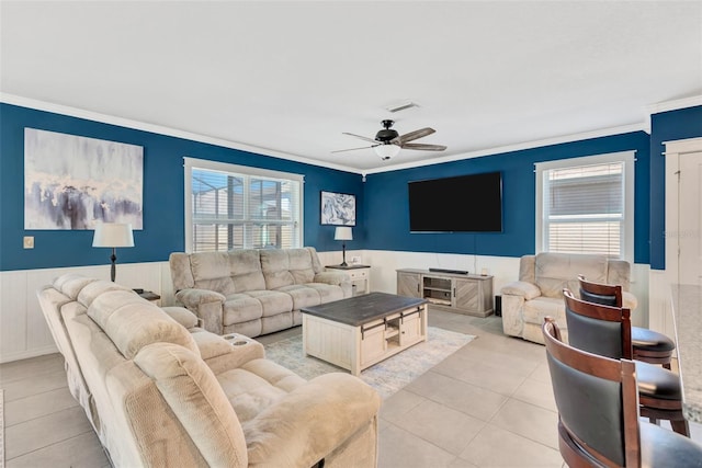 living room with crown molding, light tile patterned floors, ceiling fan, visible vents, and a wainscoted wall