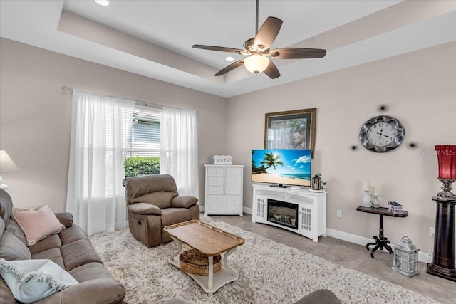 living room featuring baseboards, a ceiling fan, a raised ceiling, and a glass covered fireplace