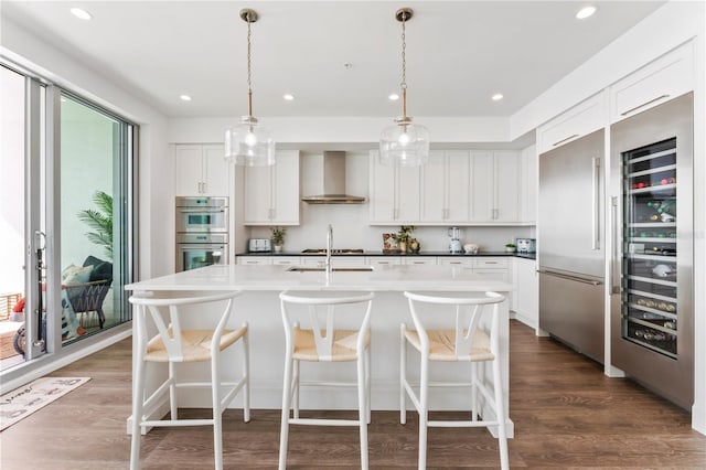 kitchen with wall chimney range hood, stainless steel appliances, white cabinetry, and an island with sink