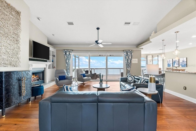 living area with wood finished floors, a warm lit fireplace, vaulted ceiling, ceiling fan, and visible vents