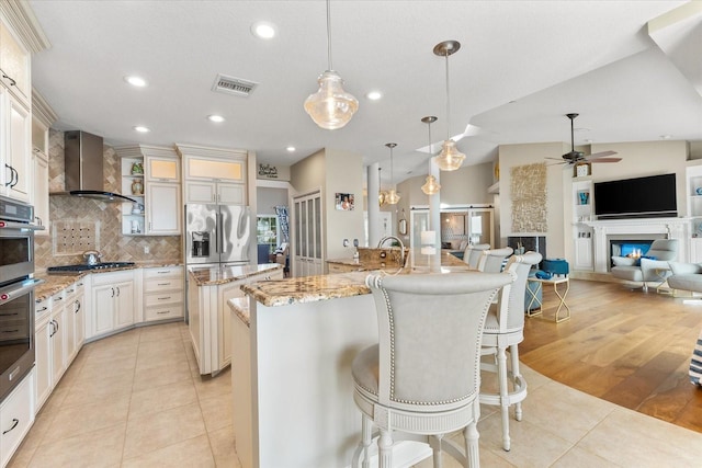 kitchen with wall chimney exhaust hood, stainless steel appliances, a large island with sink, visible vents, and backsplash