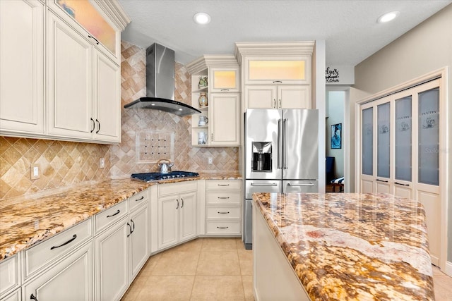 kitchen featuring light stone counters, wall chimney range hood, glass insert cabinets, tasteful backsplash, and appliances with stainless steel finishes