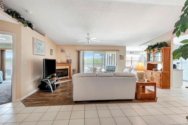 living room with light tile patterned floors, a textured ceiling, ceiling fan, baseboards, and a glass covered fireplace