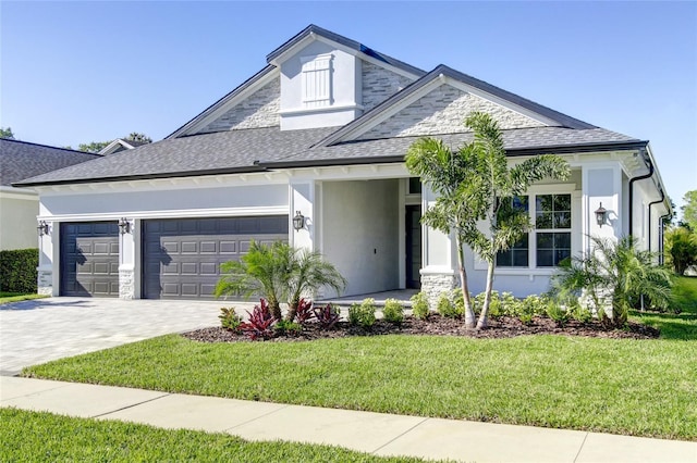 view of front of property featuring a front yard, roof with shingles, decorative driveway, an attached garage, and stone siding