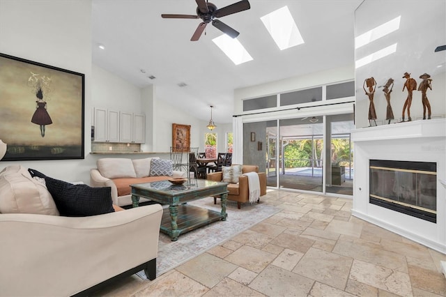 living area with stone tile floors, a glass covered fireplace, a skylight, ceiling fan, and high vaulted ceiling