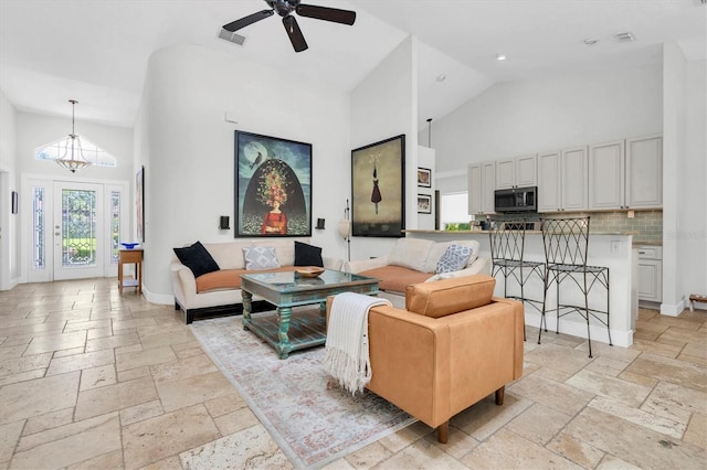 living room featuring high vaulted ceiling, baseboards, stone tile flooring, and ceiling fan with notable chandelier