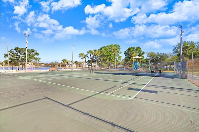 view of tennis court with fence, playground community, and a water view