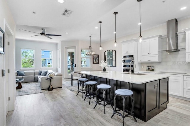 kitchen with a sink, black electric stovetop, wall chimney exhaust hood, visible vents, and ceiling fan with notable chandelier