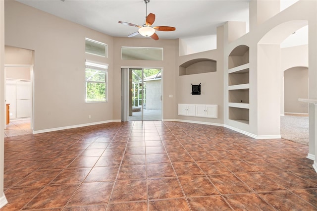 unfurnished living room featuring tile patterned floors, built in shelves, ceiling fan, arched walkways, and baseboards