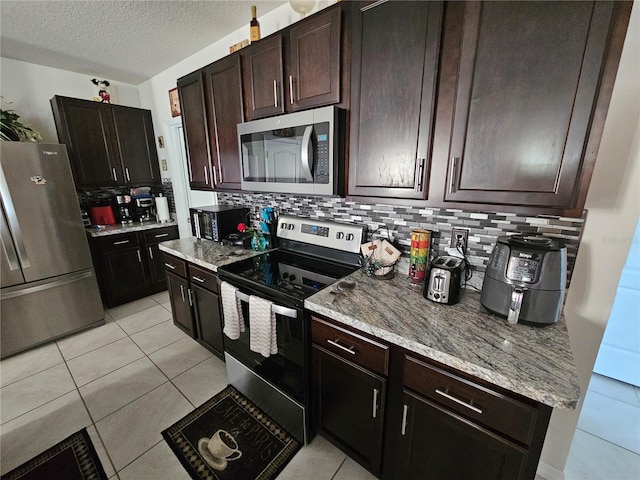 kitchen featuring decorative backsplash, light tile patterned floors, dark brown cabinetry, stainless steel appliances, and light stone countertops