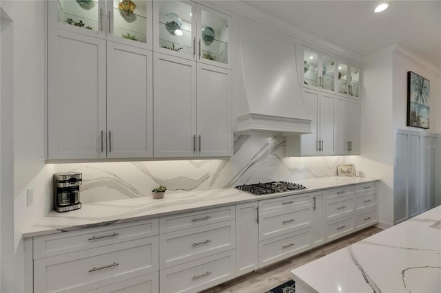 kitchen featuring light stone counters, stainless steel gas cooktop, glass insert cabinets, premium range hood, and white cabinetry