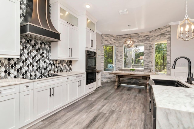 kitchen featuring an inviting chandelier, backsplash, light stone counters, premium range hood, and dobule oven black