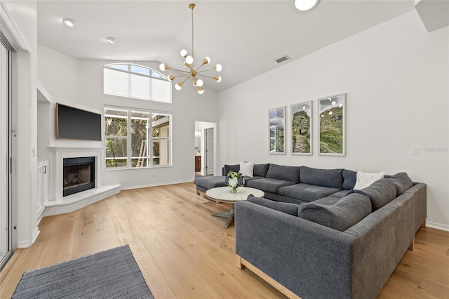 living area featuring high vaulted ceiling, a fireplace with raised hearth, light wood-style flooring, a notable chandelier, and visible vents