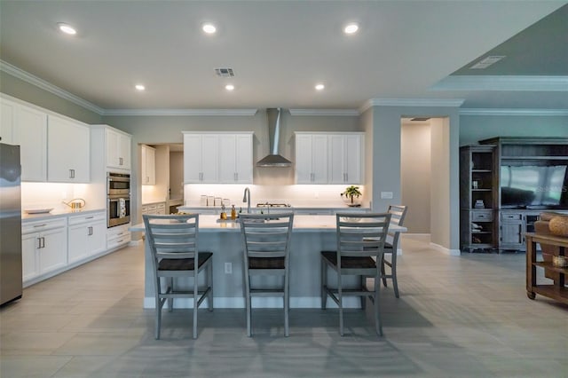 kitchen with wall chimney range hood, a kitchen breakfast bar, a kitchen island with sink, stainless steel appliances, and visible vents