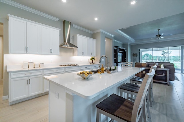 kitchen with crown molding, a center island with sink, a kitchen bar, white cabinets, and wall chimney exhaust hood