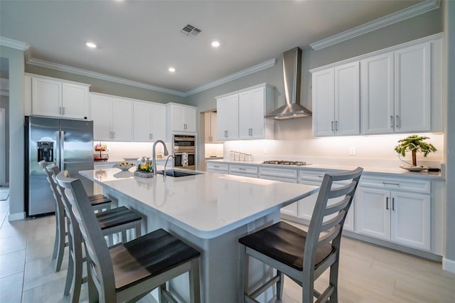 kitchen with wall chimney exhaust hood, stainless steel appliances, white cabinets, a center island with sink, and ornamental molding