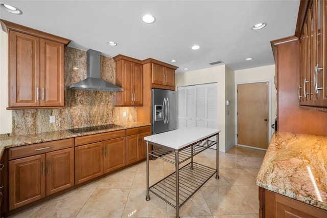 kitchen featuring brown cabinetry, decorative backsplash, stainless steel fridge with ice dispenser, black electric cooktop, and wall chimney exhaust hood