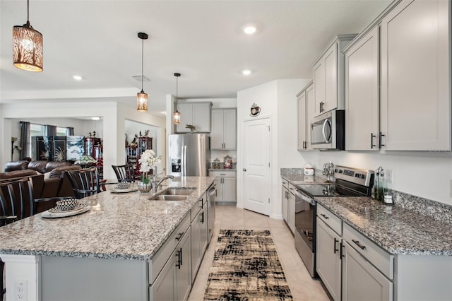 kitchen with a sink, a kitchen island with sink, gray cabinets, stainless steel appliances, and light stone countertops