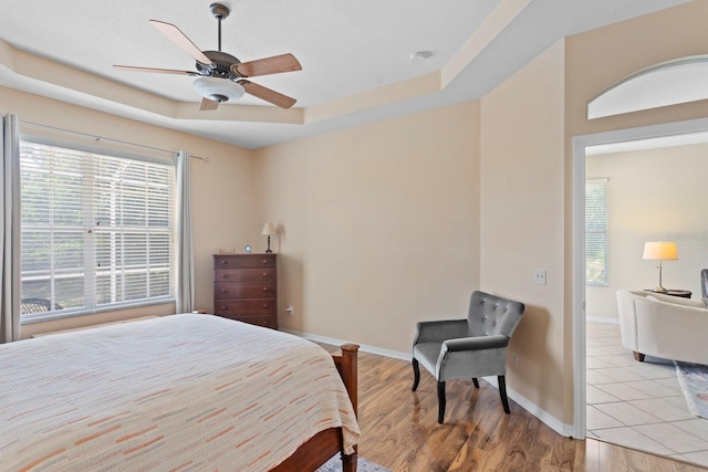 bedroom featuring ceiling fan, light wood-style flooring, a raised ceiling, and baseboards