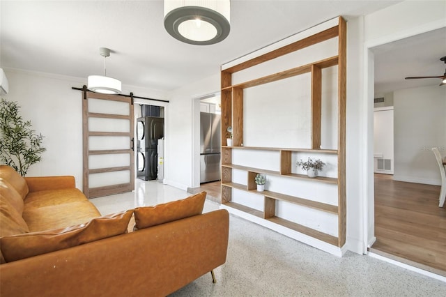 living area with stacked washer and clothes dryer, a barn door, a ceiling fan, baseboards, and dark speckled floor