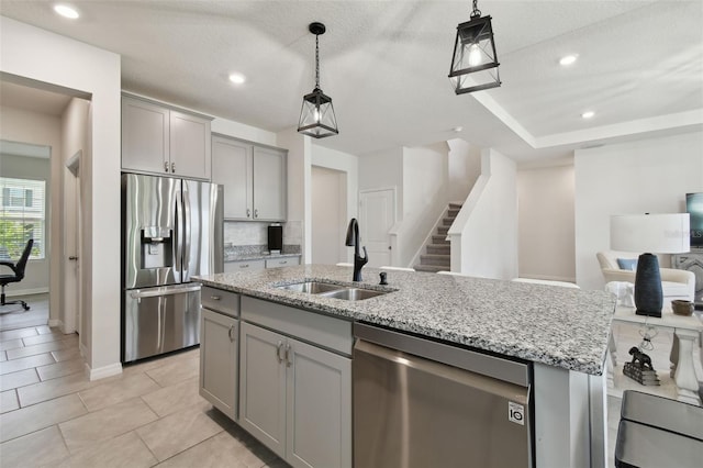 kitchen featuring a kitchen island with sink, appliances with stainless steel finishes, gray cabinetry, light stone counters, and a sink