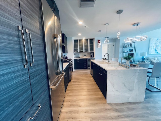 kitchen featuring a sink, a kitchen island with sink, dark cabinetry, hanging light fixtures, and light wood-style floors