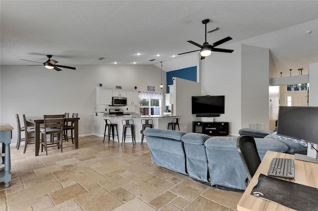 living area featuring a ceiling fan, high vaulted ceiling, a textured ceiling, stone tile floors, and baseboards
