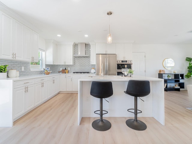 kitchen featuring wall chimney range hood, tasteful backsplash, light wood finished floors, white cabinets, and appliances with stainless steel finishes