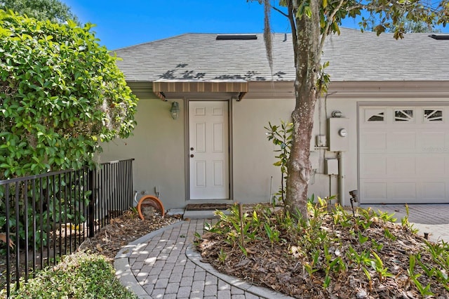 view of exterior entry featuring an attached garage, stucco siding, roof with shingles, and fence