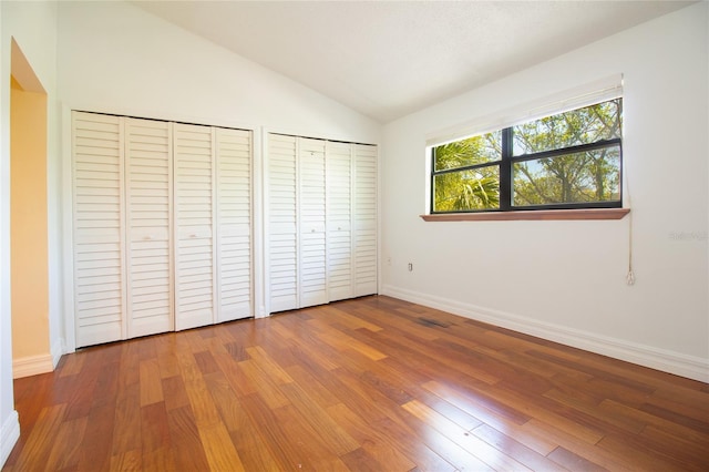 unfurnished bedroom with two closets, lofted ceiling, wood finished floors, and baseboards