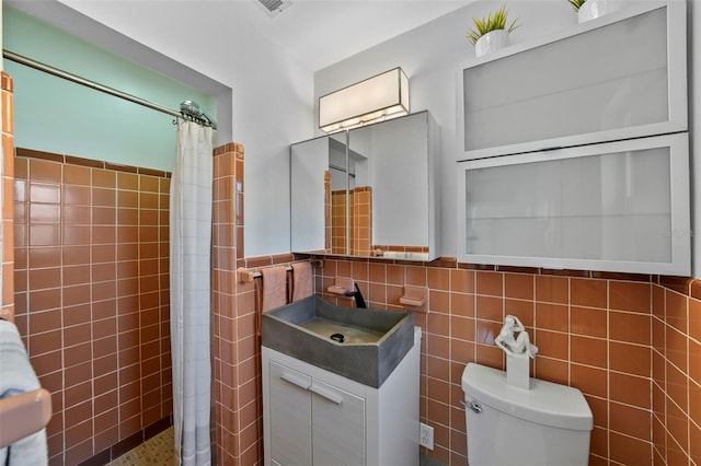 bathroom with tile walls, vanity, toilet, a tile shower, and wainscoting