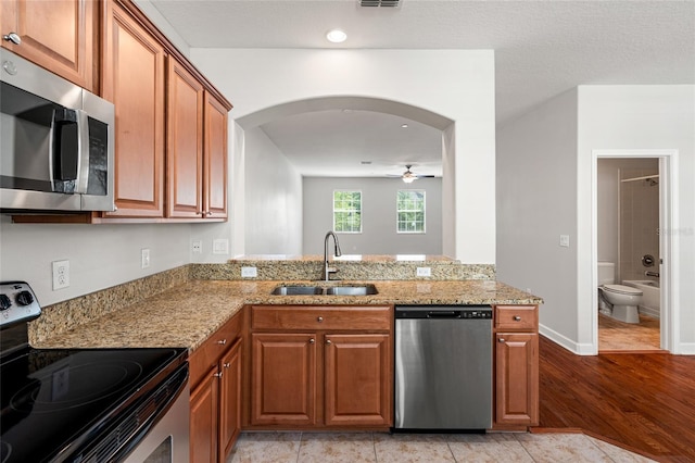 kitchen with stainless steel appliances, a sink, brown cabinets, ceiling fan, and light stone countertops