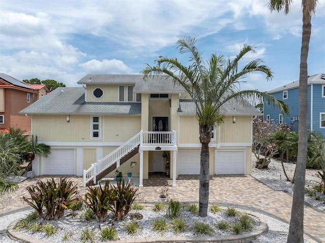 raised beach house with roof with shingles, an attached garage, decorative driveway, and stairs