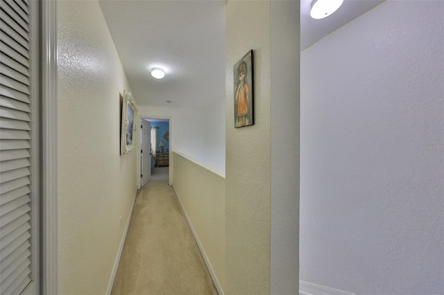 hallway featuring baseboards, light colored carpet, and a textured wall